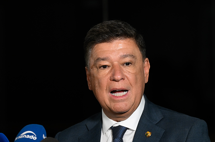 Um homem de terno fala em frente a microfones com logotipo do Senado