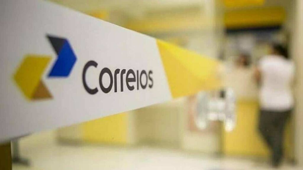 Placa com logo dos Correios em destaque dentro de unidade, com pessoas ao fundo em movimento