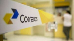 Placa com logo dos Correios em destaque dentro de unidade, com pessoas ao fundo em movimento