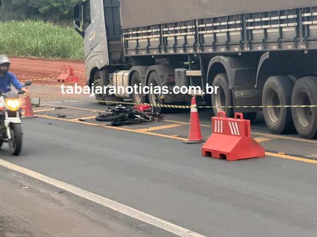 Um homem de capacete observa uma motocicleta tombada perto de um caminhão na rodovia