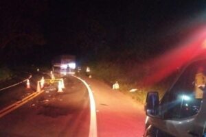 Uma estrada noturna com destroços e cones de sinalização após um acidente com um veículo parado e pessoas ao redor