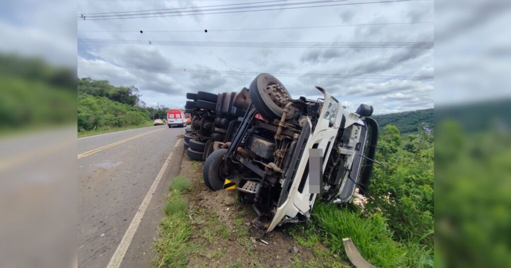 Um caminhão tombado na lateral de uma rodovia com vegetação ao redor e veículos de emergência ao fundo
