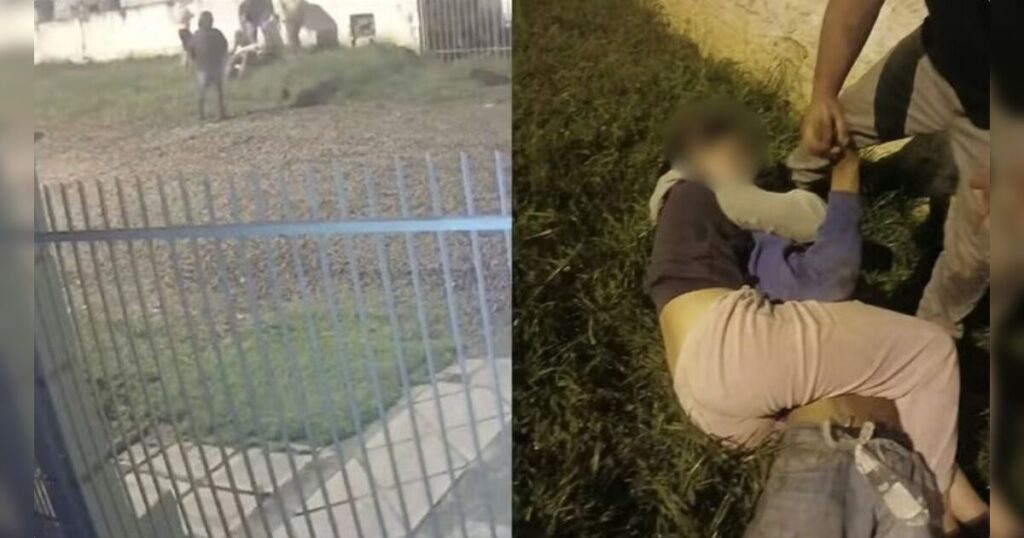 Um jovem segura um homem no chão enquanto outro observa perto de um muro com arame farpado