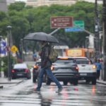 Um homem com guarda-chuva atravessa rua molhada em dia de chuva intensa com tráfego lento