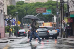Um homem com guarda-chuva atravessa rua molhada em dia de chuva intensa com tráfego lento