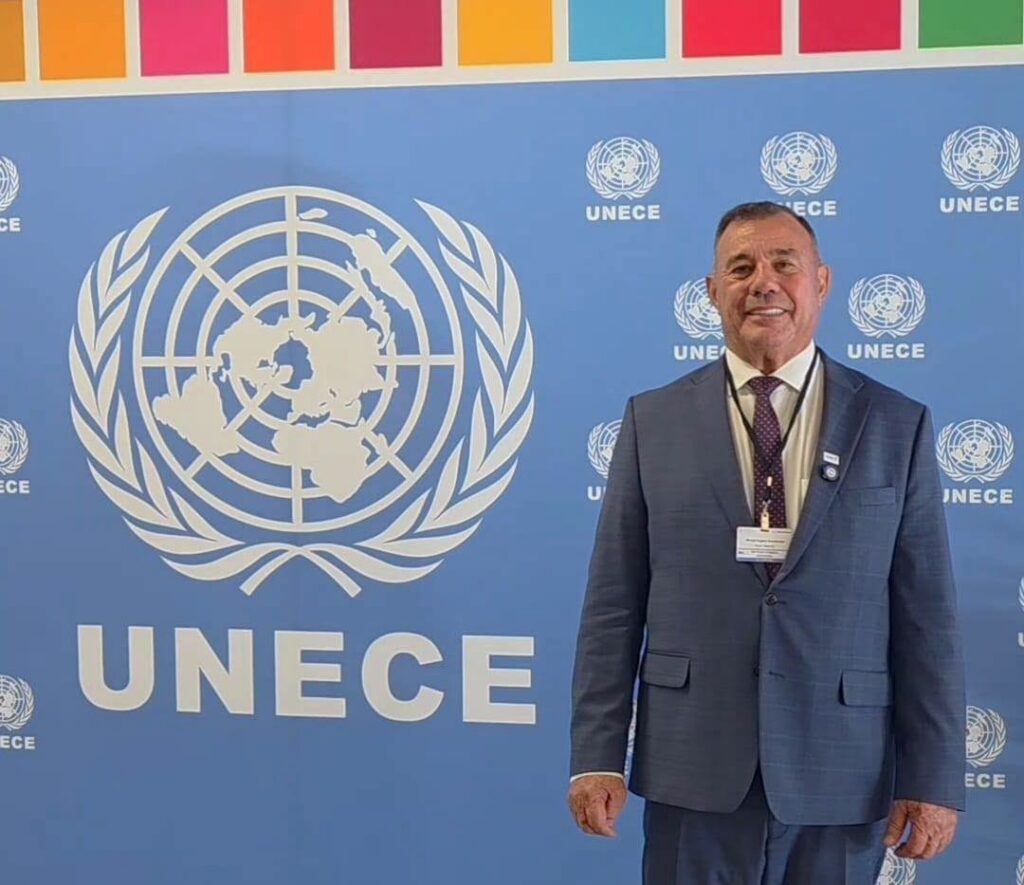 Um homem de terno posa em frente a um painel com o logo da UNECE em evento internacional