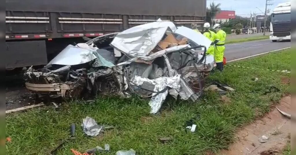 Um carro destruído após colisão com caminhão em acostamento de rodovia com dois profissionais em uniforme ao lado