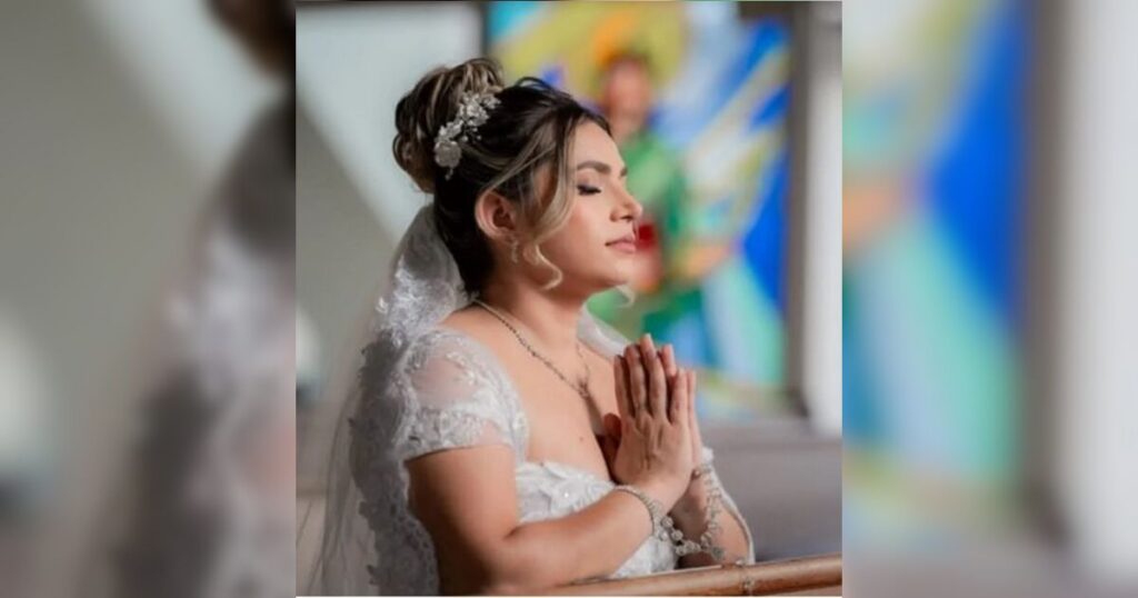 Uma mulher de vestido branco e véu com as mãos juntas em gesto de oração em um ambiente com vitrais coloridos