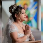 Uma mulher de vestido branco e véu com as mãos juntas em gesto de oração em um ambiente com vitrais coloridos