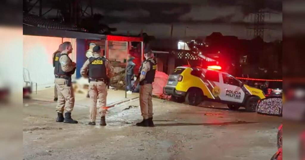 Três homens de uniforme com coletes que dizem "Polícia Militar" em área externa noturna com veículo da corporação e luzes vermelhas acesas