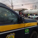 Um caminhão tanque tombado em frente a um barracão com uma viatura da Polícia Rodoviária Federal no local do acidente