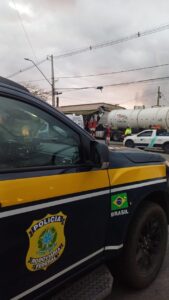 Um caminhão tanque tombado em frente a um barracão com uma viatura da Polícia Rodoviária Federal no local do acidente