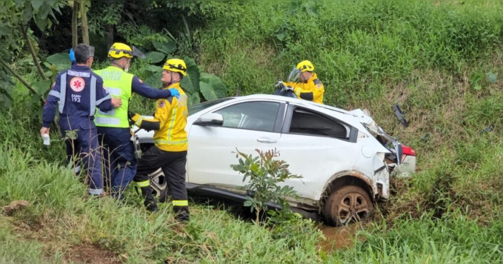 Um carro branco tombado na beira da estrada com equipes de resgate atuando ao redor