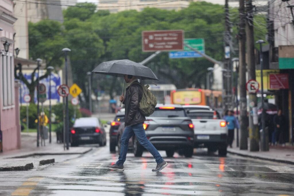 Um homem com guarda-chuva atravessa rua molhada em dia de chuva com tráfego intenso