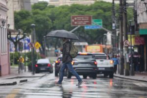 Um homem com guarda-chuva atravessa rua molhada em dia de chuva com tráfego intenso