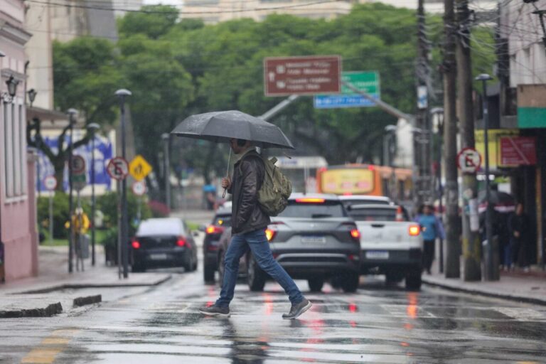 Um homem com guarda-chuva atravessa rua molhada em dia de chuva com tráfego intenso