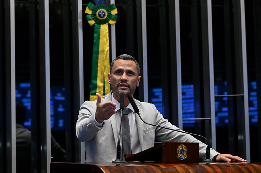 Um homem de terno fala em um púlpito com bandeira ao fundo em ambiente formal