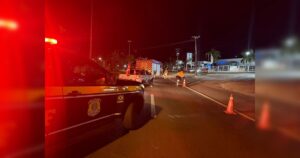 Um veículo de emergência e uma viatura policial em uma rodovia à noite com cones de sinalização e pessoas no local do acidente
