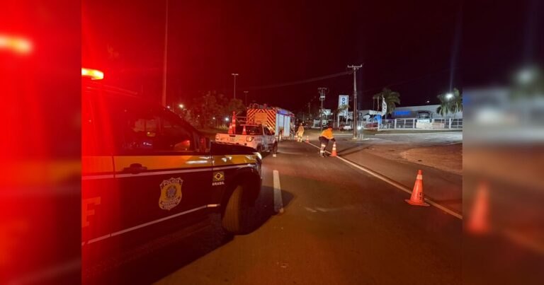 Um veículo de emergência e uma viatura policial em uma rodovia à noite com cones de sinalização e pessoas no local do acidente