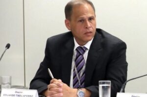Um homem de terno sentado em frente a microfone com nome na placa da mesa