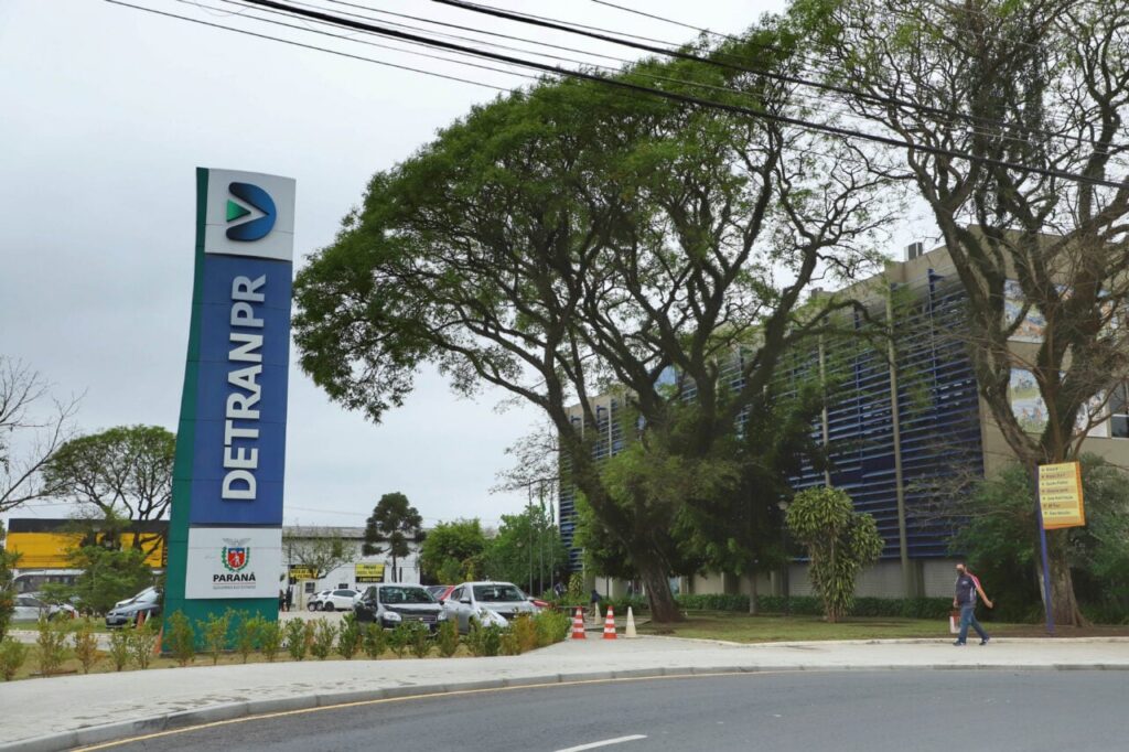 Um homem caminha em frente a um prédio com um grande letreiro de DETRAN PR ao lado de uma rua com carros estacionados