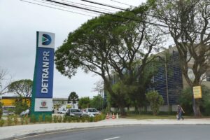 Um homem caminha em frente a um prédio com um grande letreiro de DETRAN PR ao lado de uma rua com carros estacionados