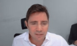 Um homem de camisa branca sentado em frente a uma câmera em ambiente interno