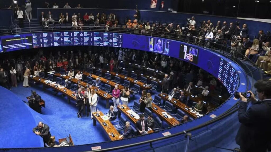 Um grupo de pessoas em um plenário com bancadas e telas ao fundo, algumas em pé e outras sentadas, em um ambiente com iluminação azul