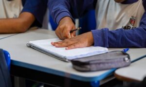 Um aluno escreve em caderno sobre carteira de sala de aula com lápis e estojo ao lado