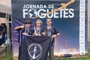 Três jovens seguram bandeira e troféu em frente a painel de foguetes