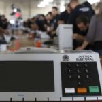 Uma urna eletrônica da Justiça Eleitoral em primeiro plano com pessoas ao fundo organizando materiais em uma mesa