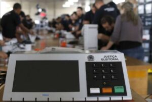 Uma urna eletrônica da Justiça Eleitoral em primeiro plano com pessoas ao fundo organizando materiais em uma mesa