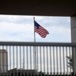 Bandeira dos Estados Unidos acena atrás de grade em edifício com estrutura metálica e concreto