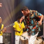 Um homem canta em um palco com músicos ao fundo usando camisetas amarelas