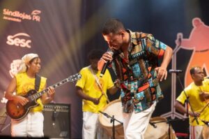 Um homem canta em um palco com músicos ao fundo usando camisetas amarelas