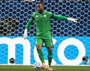 Um goleiro com uniforme verde segura a bola perto do gol em campo de futebol