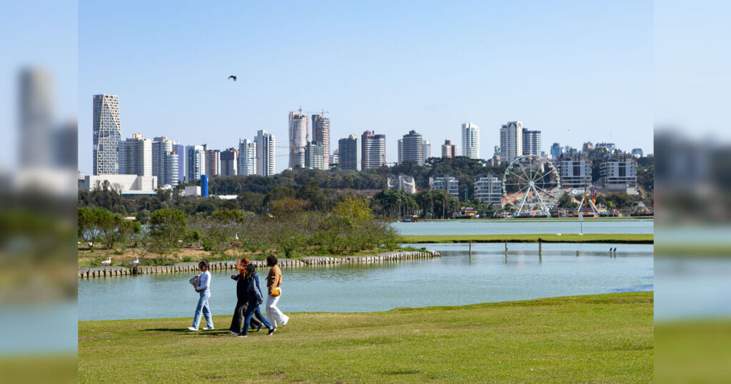 Um grupo de pessoas caminha por um gramado com uma cidade ao fundo e um lago à frente