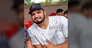 Um homem de camiseta branca e boné posa em um evento ao ar livre com outras pessoas ao fundo