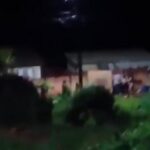 Um grupo de pessoas em frente a uma casa durante a noite com vegetação ao redor