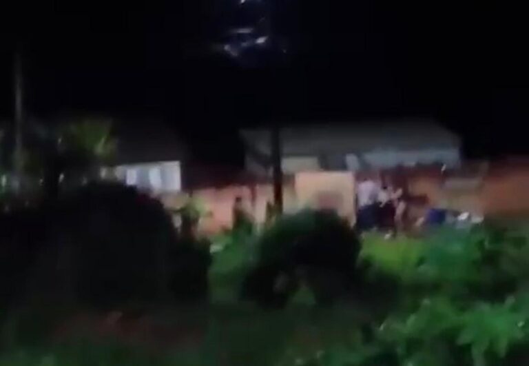 Um grupo de pessoas em frente a uma casa durante a noite com vegetação ao redor