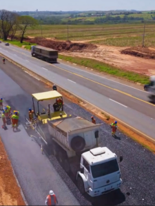 Um grupo de pessoas trabalha na pavimentação de uma rodovia com máquinas e caminhões em uma área rural