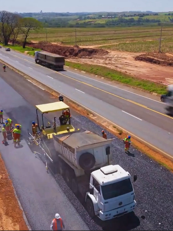 Um grupo de pessoas trabalha na pavimentação de uma rodovia com máquinas e caminhões em uma área rural