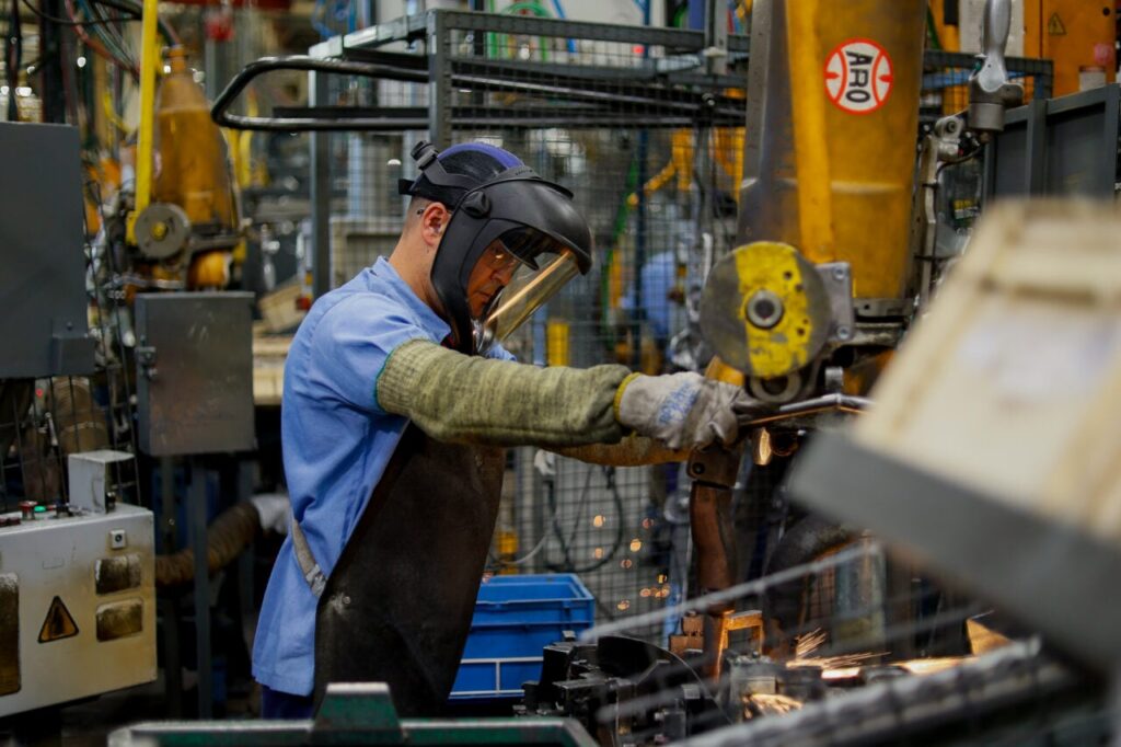 Um homem trabalha com solda em uma fábrica com equipamentos industriais ao fundo