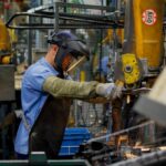 Um homem trabalha com solda em uma fábrica com equipamentos industriais ao fundo