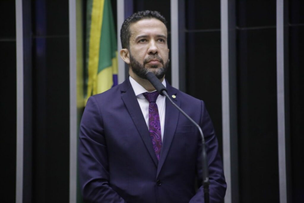Um homem de terno fala em frente a um microfone com bandeira ao fundo