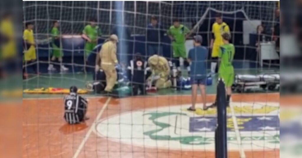 Um homem sentado no chão de uma quadra de vôlei enquanto profissionais atendem uma pessoa deitada perto do gol