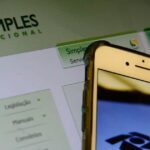 Um celular exibe o logo do Simples Nacional em frente a tela com o site do programa