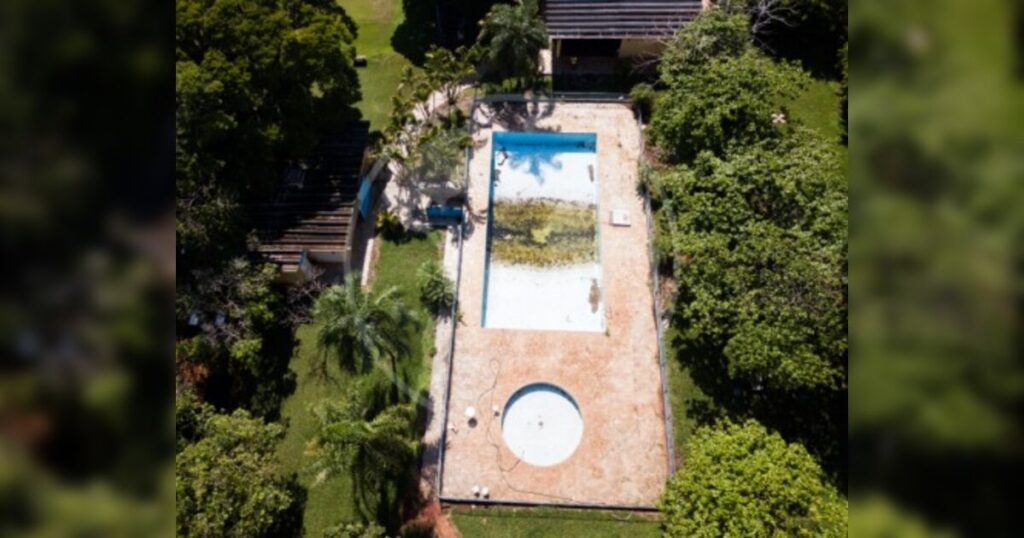 Uma piscina retangular com água parada e vegetação, cercada por gramado e árvores em área residencial