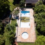 Uma piscina retangular com água parada e vegetação, cercada por gramado e árvores em área residencial