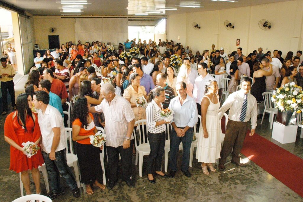 Um grupo de pessoas em cerimônia de casamento coletivo em salão com cadeiras e flores